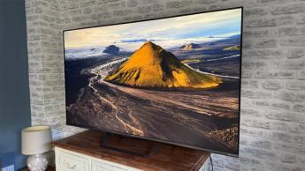 Телевизор Metz MQE7600 4K стоит на деревянной тумбе у кирпичной стены, на экране — живописный горный пейзаж