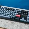 Keychron V6 mekaniskt tangentbord i svart med grå, svarta och orange tangenter visat på en stenplatta mot en blå vägg