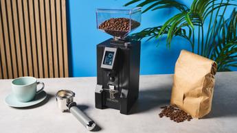 Molino de café Eureka Mignon Specialità colocado sobre una encimera de cocina junto a una pared azul, mostrando su diseño elegante y capacidades de preparación precisas.