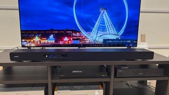 Саундбар Yamaha True X Surround 90A с тыловыми колонками и сабвуфером, установленный в простой комнате с телевизором