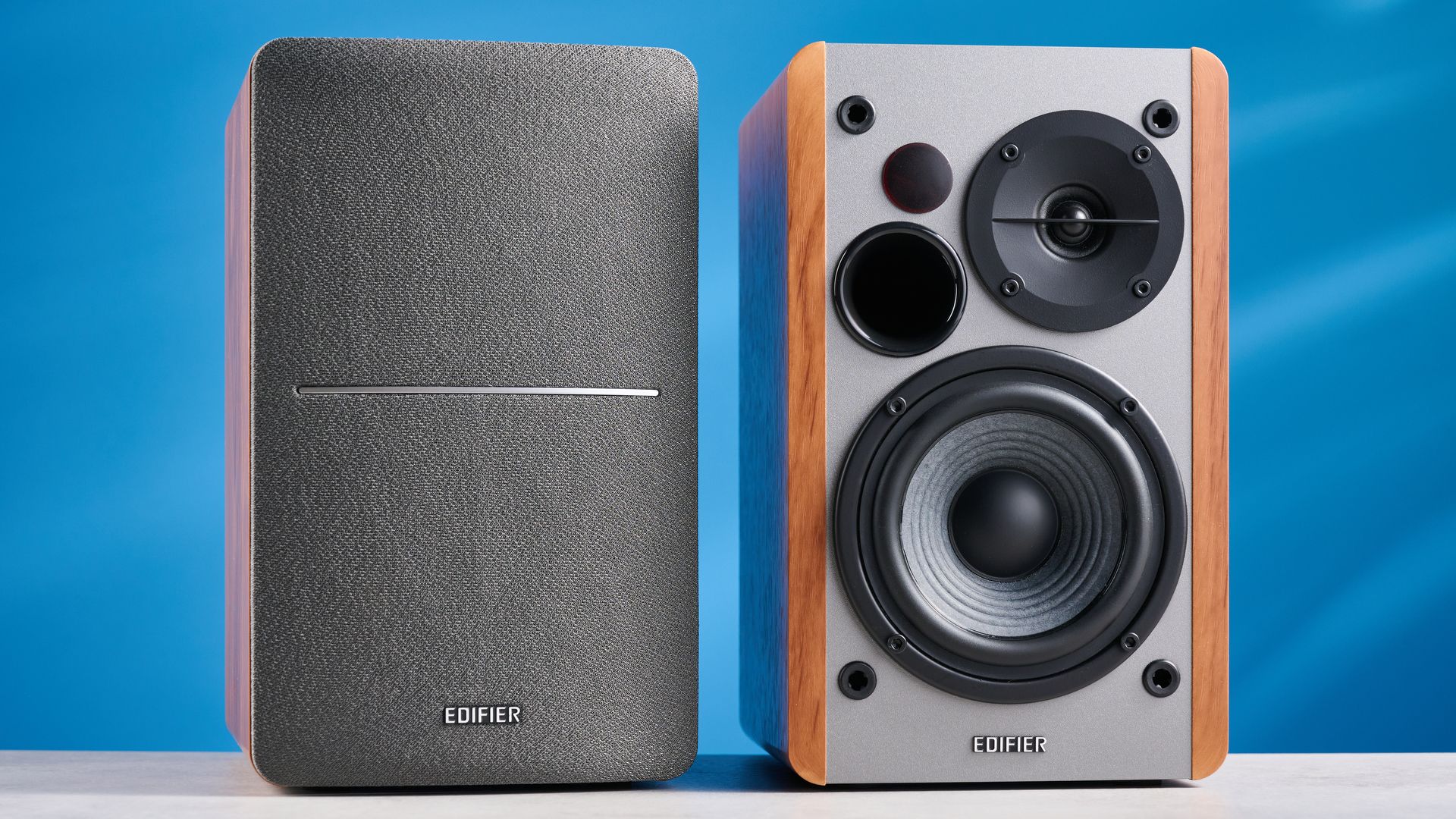 Die Edifier R1280T Regallautsprecher in Holzbraun vor blauem Hintergrund, mit Fokus auf Lautstärkeregler, Lautsprechertreiber, Hochtöner, RCA- und Aux-Eingänge sowie Fernbedienung