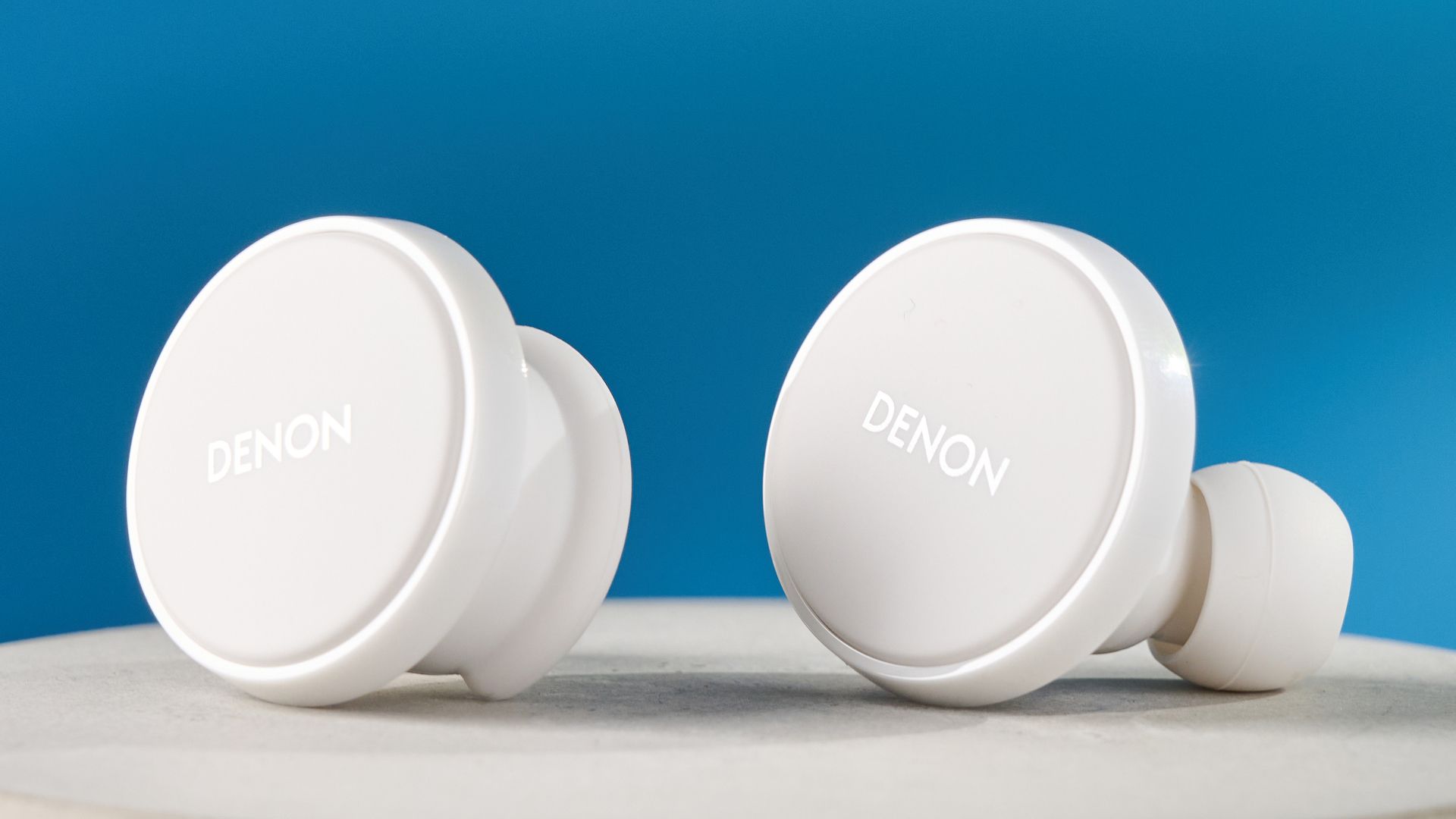 Weiße Denon perL Pro True-Wireless-Kopfhörer mit runden Ohrstöpseln und passender weißer Ladehülle vor blauem Hintergrund