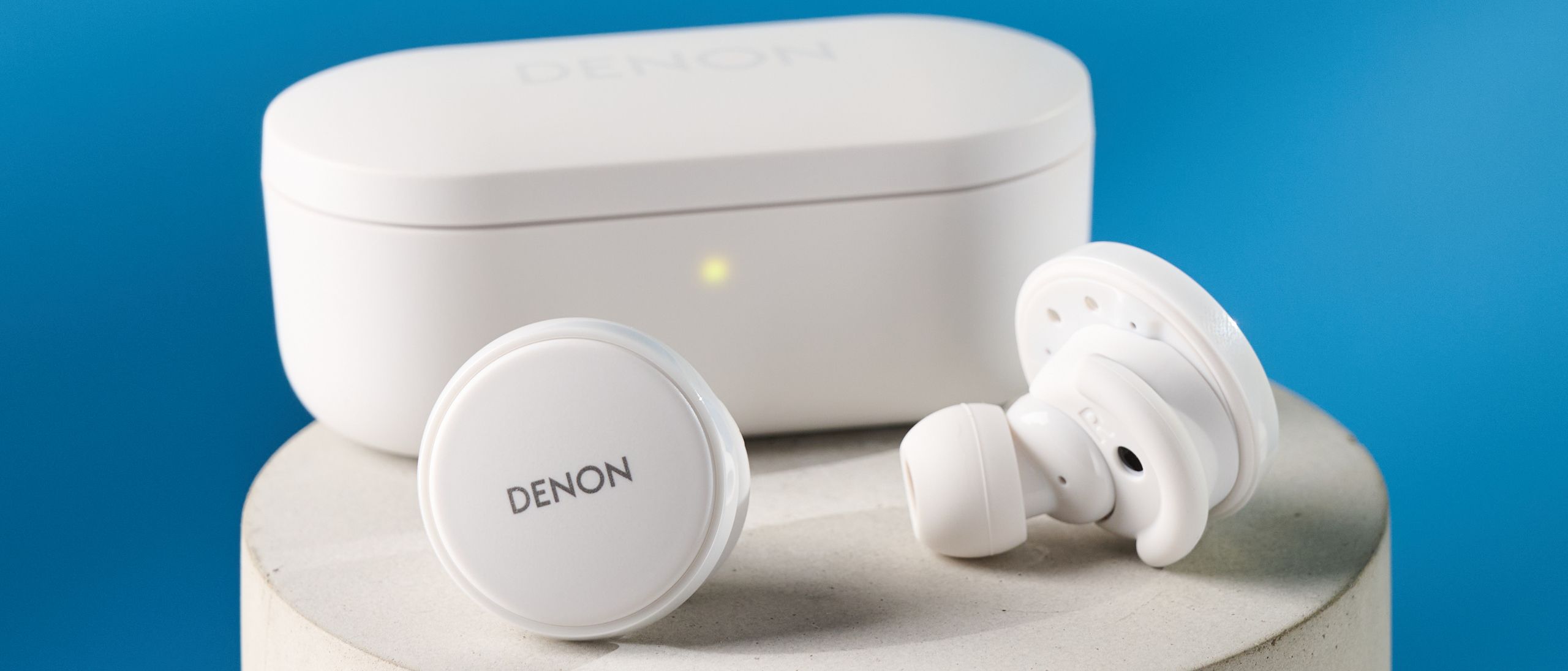 Weiße Denon perL Pro kabellose Ohrhörer mit runden Earbuds und passender weißer Ladebox auf blauem Hintergrund