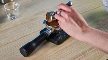La macchina per espresso Breville Bambino Plus in acciaio inox con accessori inclusi, come bricco per il latte, pressino e portafiltro, mostrata su sfondo blu mentre estrae l'espresso