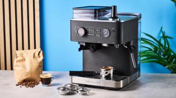 Uma máquina de espresso preta da Kitchenaid com botões cromados, manômetro de pressão e vaporizador de leite, exibida junto a um copo de espresso prensado e um pacote de grãos de café inteiros, sobre um fundo azul