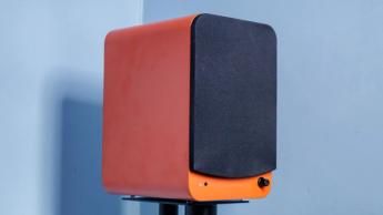 Ein orangefarbener Kanto Ren Bluetooth-Lautsprecher mit Frontgitter, platziert vor einer blauen Wand