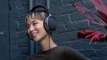 En ung person med et androgynt udseende iført Audeze LCD-S20 planmagnetiske hovedtelefoner, stående foran en mørkegrå murstensvæg og en mørkerød plante