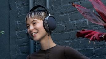 En ung person med et androgynt udseende iført Audeze LCD-S20 planmagnetiske hovedtelefoner, stående foran en mørkegrå murstensvæg og en mørkerød plante