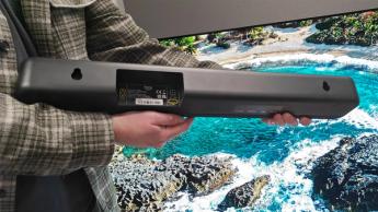 Обозреватель держит саундбар Amazon Fire TV перед телевизором с изображением морского побережья, видны разъёмы подключения саундбара