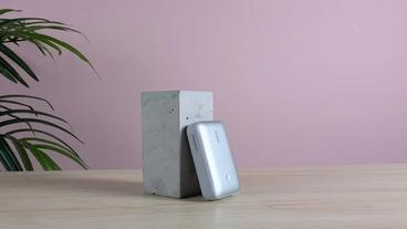 Cargador portátil Aukey Spark Mini 20000 descansando sobre un pedestal en un escritorio, con una pared rosa y una planta verde al fondo