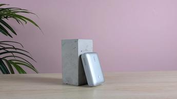 Cargador portátil Aukey Spark Mini 20000 descansando sobre un pedestal en un escritorio, con una pared rosa y una planta verde al fondo
