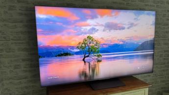 65-дюймовый 4K ЖК-телевизор TCL на деревянной тумбе перед кирпичной стеной, на экране — живописное озеро и дерево