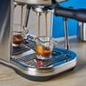 A máquina de espresso Breville Bambino Plus em aço inoxidável prateado sobre um fundo azul, com acessórios incluindo jarra de leite, prensador e porta-filtro, durante o preparo de um espresso