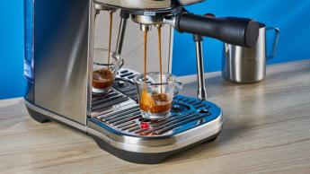 A máquina de espresso Breville Bambino Plus em aço inoxidável prateado sobre um fundo azul, com acessórios incluindo jarra de leite, prensador e porta-filtro, durante o preparo de um espresso
