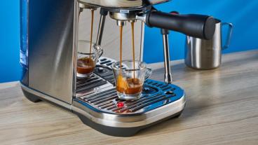 La macchina per espresso Breville Bambino Plus in acciaio inox color argento su sfondo blu, con accessori inclusi come lattiera, pressino e portafiltro, mostrata mentre eroga un espresso
