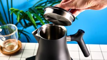 La tetera eléctrica Fellow Corvo EKG en negro mate sobre un fondo azul, acompañada de una taza de vidrio y una mesa en blanco y negro