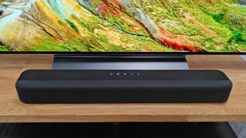 Звуковая панель Amazon Fire TV на деревянной тумбе под телевизором перед экраном с изображением горного пейзажа