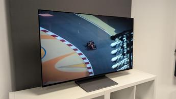 Телевизор Samsung S90F OLED 48 дюймов на белой деревянной тумбе, на экране — вид сверху на болид Формулы-1, мчащийся по трассе