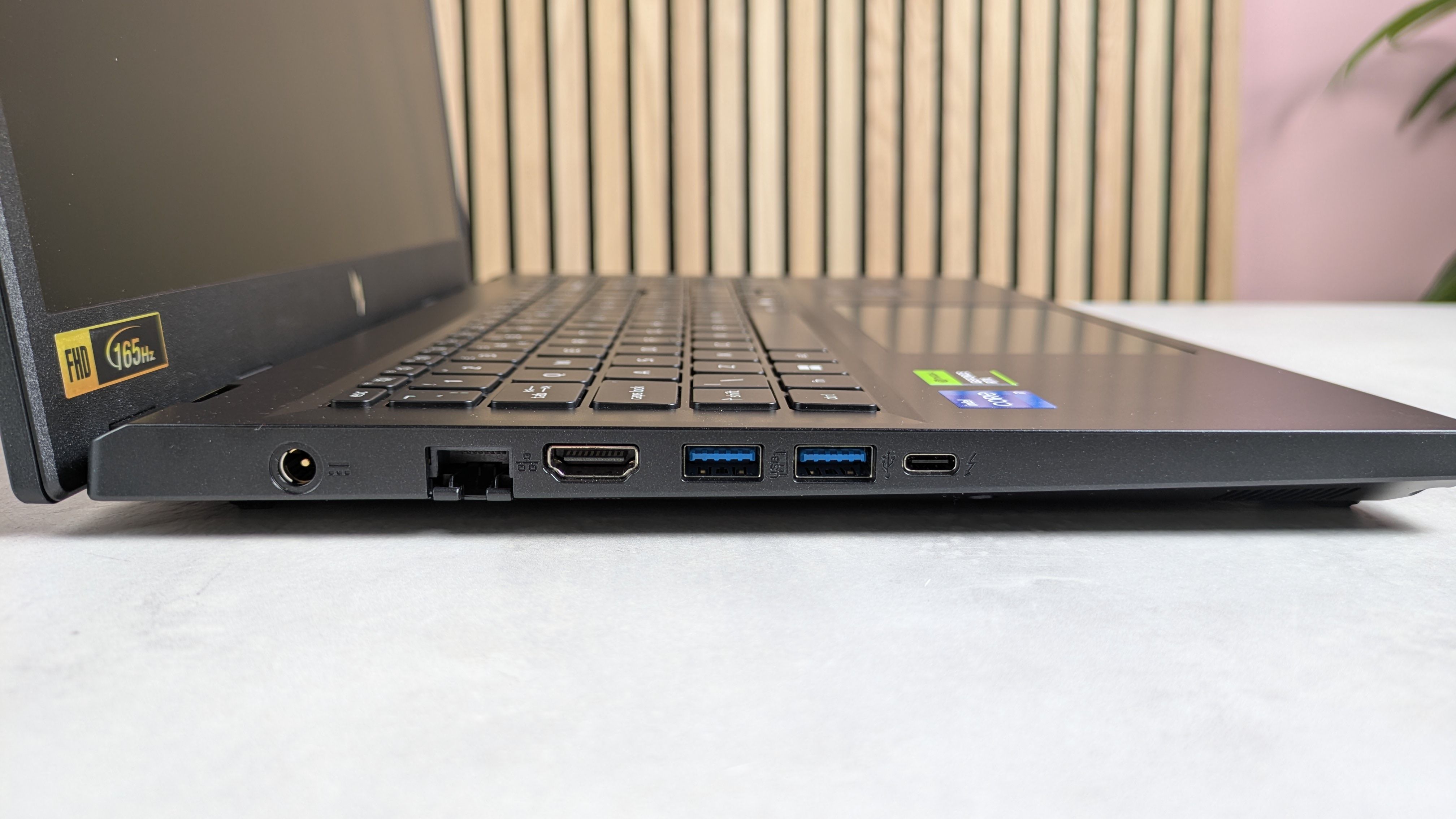 木製パネルのデスク上に置かれたAcer Nitro V 15ノートパソコンの左側ポート。背後には観葉植物とピンク色の壁が見える。