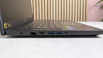 Acer Nitro V 15-bärbar dator, vänster sida med portar, på ett skrivbord med träpaneler, grön växt och rosa vägg i bakgrunden.