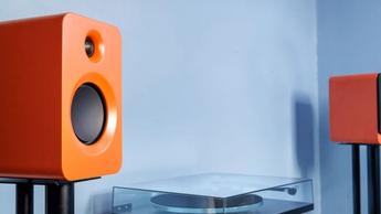 Ein Paar orangefarbene Kanto Ren Lautsprecher auf Ständern mit einem Plattenspieler dazwischen vor einer blauen Wand