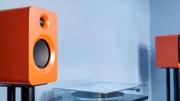 Ein Paar orangefarbene Kanto Ren Lautsprecher auf Ständern mit einem Plattenspieler dazwischen vor einer blauen Wand