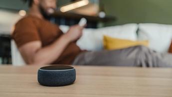 Giovane uomo afroamericano che si rilassa sul divano e naviga sul suo smartphone, con uno smart speaker sul tavolino del soggiorno