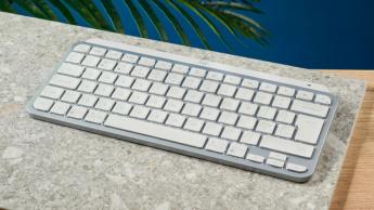 밝은 회색의 로지텍 MX Keys Mini 무선 키보드가 돌판 위에 놓여 있고, 파란색 벽을 배경으로 하고 있습니다.