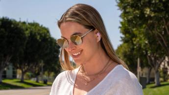 En ung kvinde med solbriller lytter til musik på JBuds Pods, mens hun kigger på sin telefon på en allé med træer