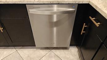 Sebuah mesin pencuci piring Whirlpool berbahan stainless steel terpasang di antara lemari dapur hitam dengan meja marmer dan lantai keramik.