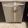 Sebuah mesin pencuci piring Whirlpool berbahan stainless steel terpasang di antara lemari dapur hitam dengan meja marmer dan lantai keramik.