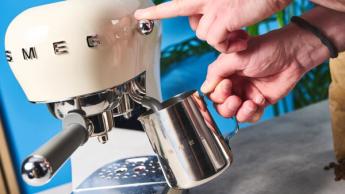 Uma máquina de espresso retrô Smeg cor creme com um pacote de café ao lado, sobre um fundo azul