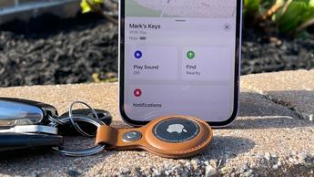 AirTag de Apple sujeto a un llavero de cuero, colocado sobre una pared junto a un iPhone que muestra la app Encontrar