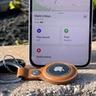 Apple AirTag dalam gantungan kunci kulit cokelat yang tergantung di dinding, di samping iPhone yang menampilkan aplikasi Lacak Saya