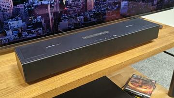 Саундбар Sharp HT-SB700 с поддержкой Dolby Atmos на деревянной стойке перед телевизором, на экране которого изображён городской пейзаж