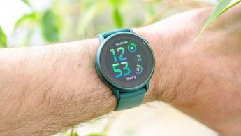 Primer plano de un smartwatch Garmin Vivoactive 6 verde en la muñeca de una persona, con follaje verde desenfocado al fondo