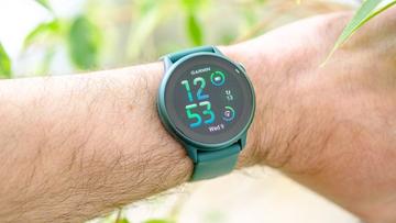 Nærbillede af et grønt Garmin Vivoactive 6 smartwatch på en persons håndled med uskarpt grønt løv i baggrunden