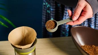 Cafeteira pour-over Wacaco Cuppamoka de aço inoxidável com filtros de papel marrons e tampa de bico, sobre fundo azul