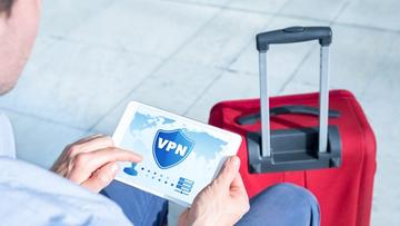 En man använder en VPN på sin laptop på en flygplats med röd resväska bredvid sig