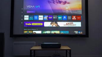 Ультракороткофокусный проектор Hisense PL2 на деревянной тумбе для AV-аппаратуры отображает главный экран ОС VIDAA в тестовой комнате