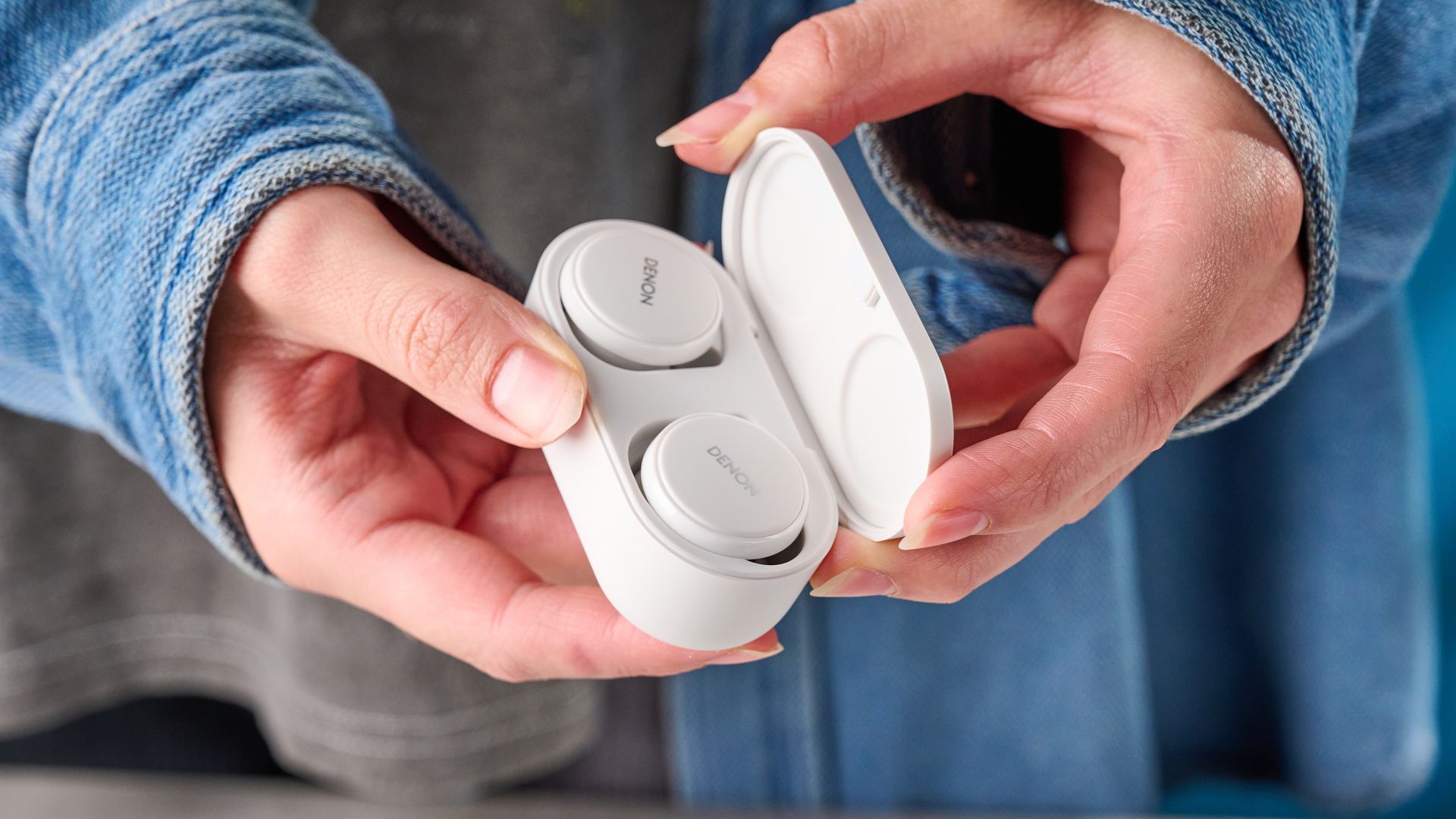 Die Denon perL Pro kabellosen Ohrhörer in Weiß mit runden Ohrstöpseln und passendem weißen Ladecase vor blauem Hintergrund