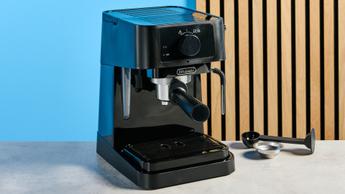A máquina de espresso De'Longhi Stilosa preta com porta-filtro, seletor para preparo de espresso e vaporização de leite, além de socador de plástico, exibida sobre um fundo azul