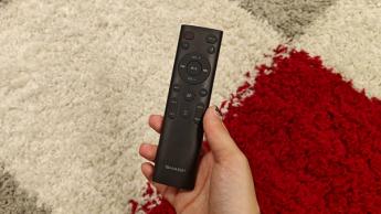 Рука держит пульт от саундбара Sharp HT-SB700 с поддержкой Dolby Atmos над ковром с красным, белым и серым узором