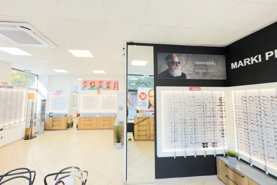 Szerokie ujęcie sali sprzedaży z lustrem i czarną ścianą marek premium z oprawami okularowymi w salonie VisionOptyk we Wrześni