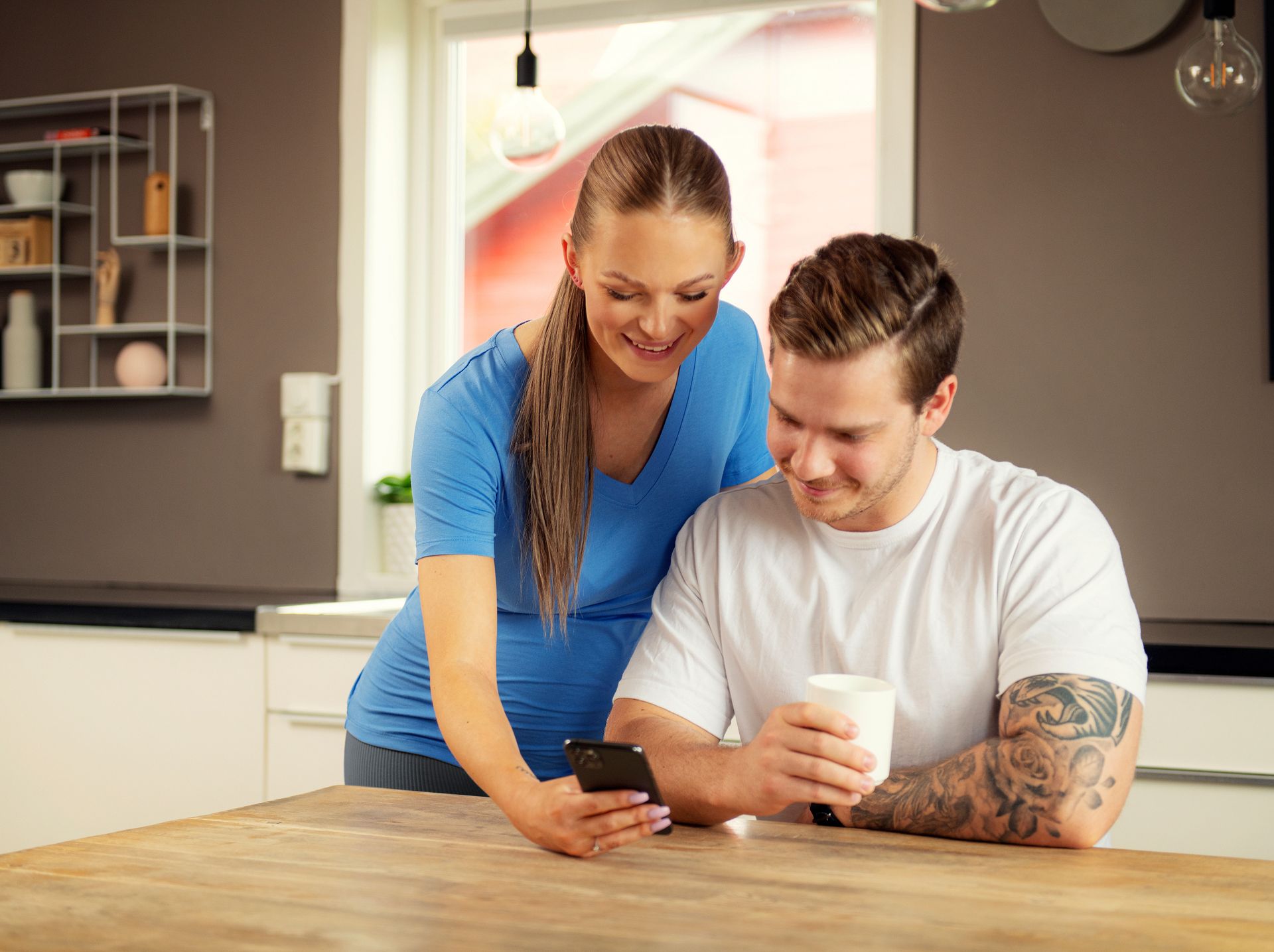 Foto av ungt par ved et bord. Hun står og viser noe på telefonen til en mann som sitter og holder en kaffekopp.