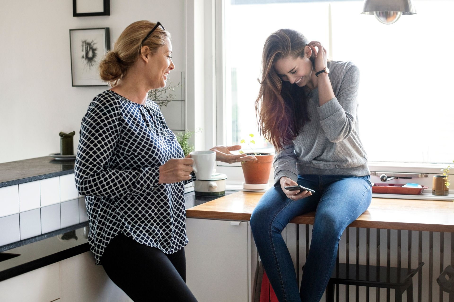 Foto av mor og datter som prater ved kjøkkenbenken. Moren lener seg inntil kjøkkenbenken og ser på datteren. Hun sitter på et bord og ser på mobiltelefonen hun har i hånden.