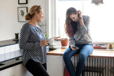 Foto av mor og datter som prater ved kjøkkenbenken. Moren lener seg inntil kjøkkenbenken og ser på datteren. Hun sitter på et bord og ser på mobiltelefonen hun har i hånden.