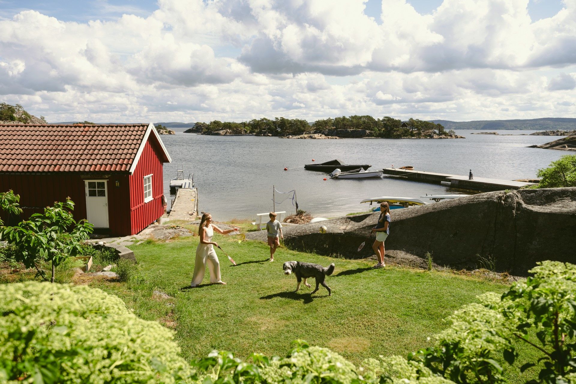 Mor, to barn og hund spiller badminton på en gressplen ved sjøen. Rødt naust og båter i bakgrunnen. Foto.
