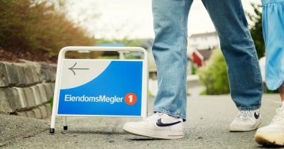Foto av et visningsskilt der det står EiendomsMegler 1. Ved siden av ser vi bena på to personer.