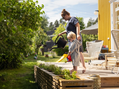 Foto av en dame og et barn foran et gult hus. De står på en terrassen og vanner grønne planter fra hver sin kanne.
